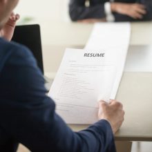 Back view of employer reading resume focus on document, job applicant waiting for hiring decision at desk on background. Hiring manager studying long list of candidates achievements and experiences