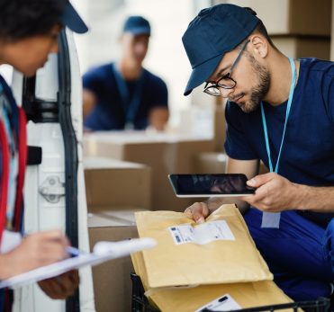Young delivery man using digital tablet for scanning bar code on package label while preparing for shipment with his coworker.