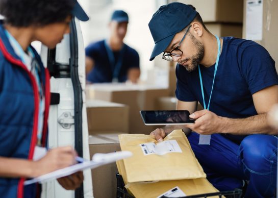 Young delivery man using digital tablet for scanning bar code on package label while preparing for shipment with his coworker.
