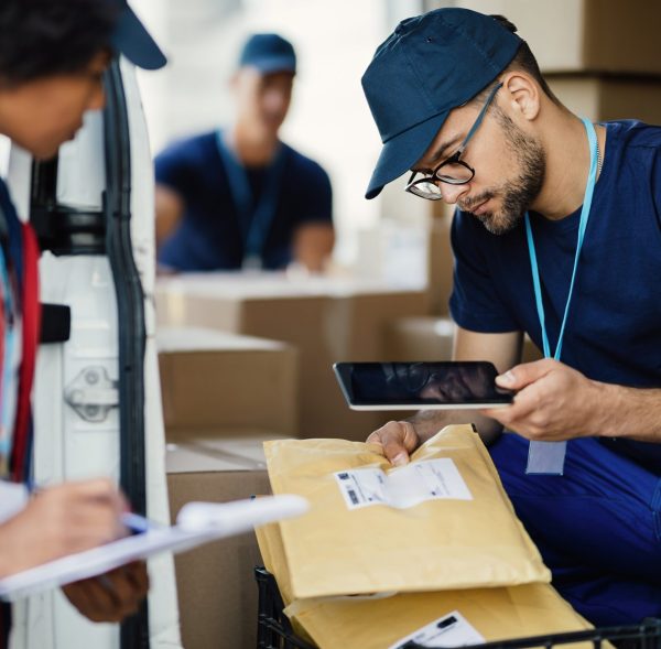Young delivery man using digital tablet for scanning bar code on package label while preparing for shipment with his coworker.