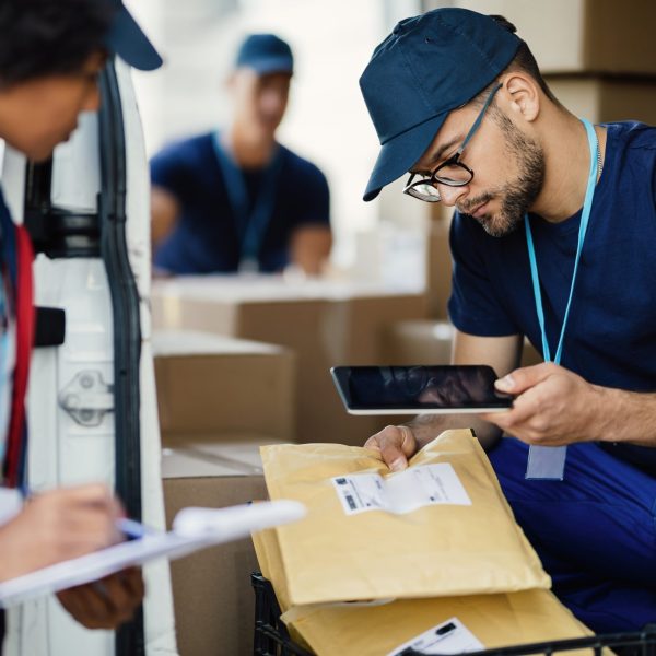 Young delivery man using digital tablet for scanning bar code on package label while preparing for shipment with his coworker.