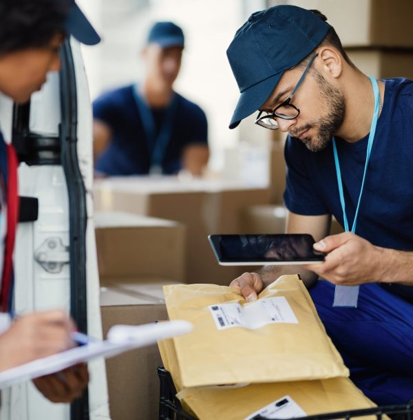 Young delivery man using digital tablet for scanning bar code on package label while preparing for shipment with his coworker.