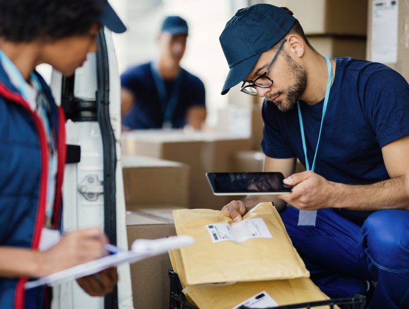 Young delivery man using digital tablet for scanning bar code on package label while preparing for shipment with his coworker.