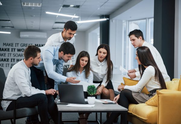 Guy shows document to a girl. Group of young freelancers in the office have conversation and working.